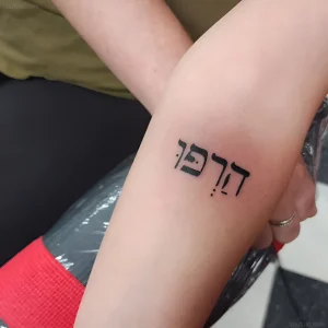Фото татуировки: черная надпись на иврите «Дерфек» на внутренней стороне предплечья, выполненная четкими линиями.