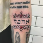 Фото татуировки на руке: ивритская надпись "אלהבה" (Ahavah - любовь) обрамлена стилизованными цветами и листьями в традиционной технике.