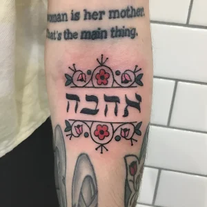 Фото татуировки на руке: ивритская надпись "אלהבה" (Ahavah - любовь) обрамлена стилизованными цветами и листьями в традиционной технике.