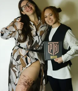 Дві жінки стоять, одна з яких тримає нагороду, а інша демонструє татуювання на нозі.