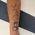 Черно-белая татуировка на руке: чашка кофе, молекула кофеина и кофейное пятно, символизирующие любовь к напитку и его воздействие.
