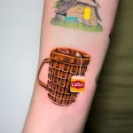Фото татуировки: реалистичная чашка горячего напитка с пакетиком чая Lipton и изображением уютного домика на заднем плане.