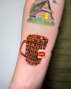 Фото татуировки: реалистичная чашка горячего напитка с пакетиком чая Lipton и изображением уютного домика на заднем плане.
