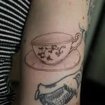 Черно-белая татуировка чашки с блюдцем, украшенная витиеватыми растительными элементами на руке.