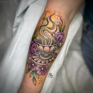 Фото татуировки чашки с паром, украшенной нежными фиолетовыми цветами на икре ноги.