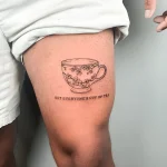 Черно-белая татуировка чашки с цветами на бедре, с надписью 'Not everyone's cup of tea'.