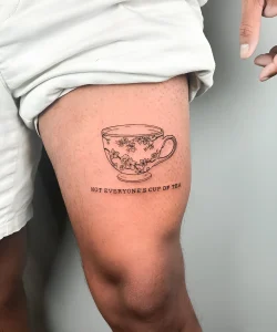 Черно-белая татуировка чашки с цветами на бедре, с надписью 'Not everyone's cup of tea'.