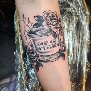 Черно-белая татуировка чашки кофе с цветами пионов и надписью 'CUP OF AMBITION' на руке