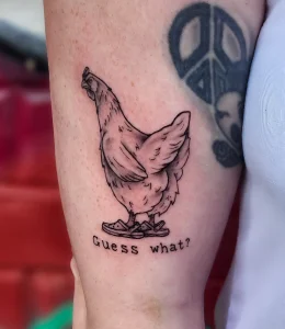 Черно-белая татуировка курицы в желтых тапочках с надписью 'Guess what?' на руке