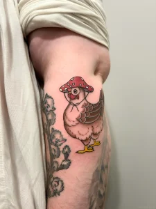 Яркая татуировка курицы в стиле олд скул с красным мухомором на бедре, окруженная растительным орнаментом.