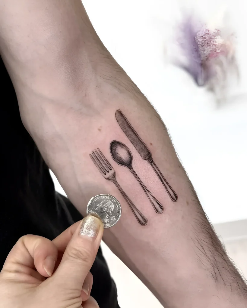 Minimalistisches Tattoo am Arm mit Löffel, Gabel, Messer und Münze, im Grafikstil.