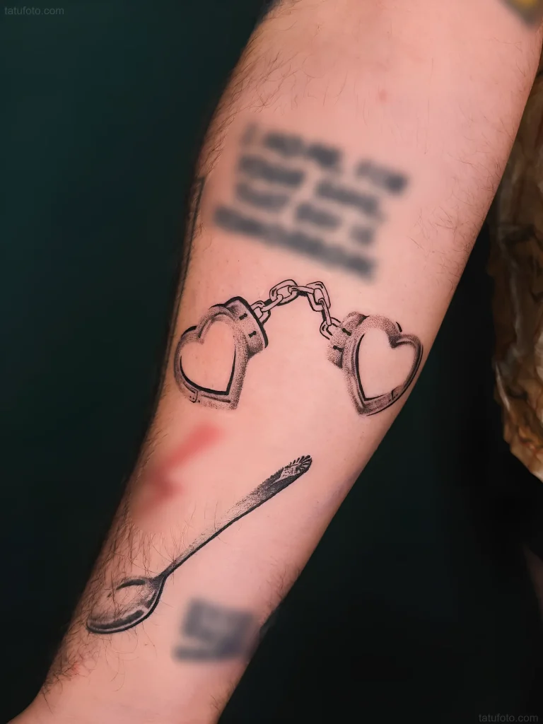 Schwarz-weißes Tattoo am Arm: ein Löffel, durchstochen von herzförmigen Handschellen, was die komplexe Verbindung von Liebe und Kontrolle symbolisiert.