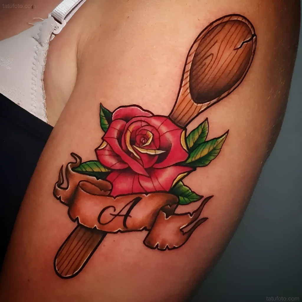 Foto eines Tattoos auf der Schulter: ein Holzlöffel, umwickelt mit einem Band mit einem Namen, und eine rote Rose im Old-School-Stil.