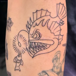 Черно-белая татуировка рыбы-удильщика на руке, выполненная в стиле олд скул с акцентом на острые зубы и светящийся приманку.