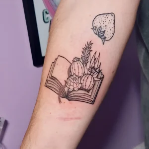 Татуировка в виде раскрытой книги, из которой прорастают суккуленты и кактус, с одинокой клубникой над композицией