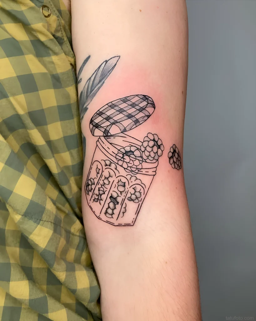 肘部上的黑白纹身，描绘了一个打开的果酱罐，里面有浆果，旁边还有几颗浆果。