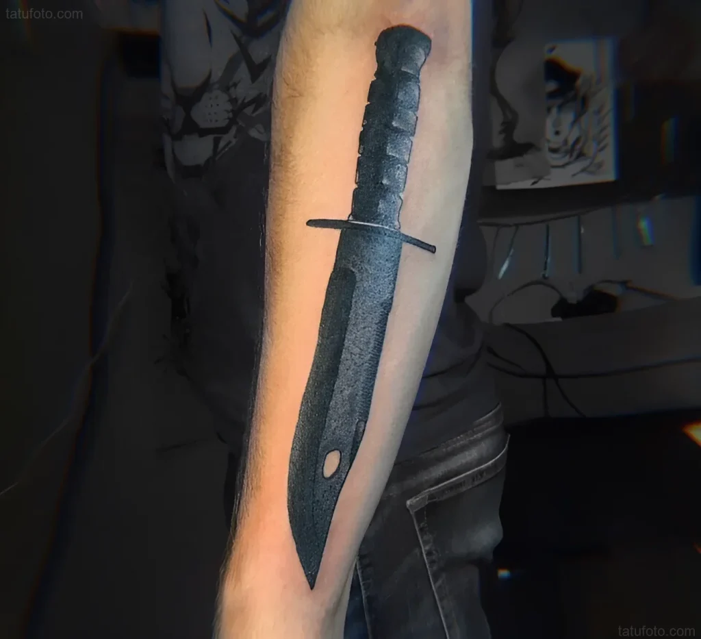 Tatouage réaliste de baïonnette sur l'avant-bras, réalisé à l'encre noire avec détail de la texture du métal.