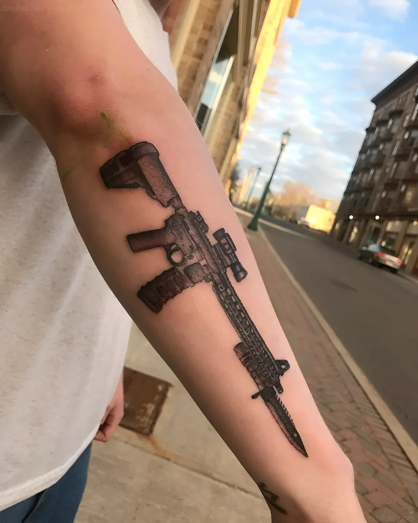 Tatouage réaliste de baïonnette attachée à un fusil d'assaut moderne, réalisé en noir et gris sur l'avant-bras.