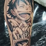 Татуировка в стиле манга: Крупный план улыбающегося лица Луффи из One Piece, держащего листовку с наградой «Wanted» с его изображением.