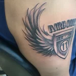 Черно-серая татуировка крыльев и щита с надписью "AIRBORNE" и числом "11" на плече