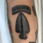 Татуировка в виде нашивки "Airborne" и стрелы ВДВ, выполненная в реалистичном стиле с использованием черных и серых оттенков.