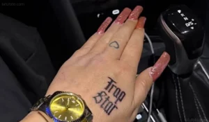 Рука Аманды Байнс с татуировкой "Trap Star" на тыльной стороне ладони и маленьким сердцем на пальце, рядом золотые часы и рычаг переключения передач автомобиля.