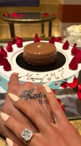 Крупный план рук с парными татуировками «Кэти» и «Ли», обручальным кольцом и тортом с надписью «Выходи за меня?».