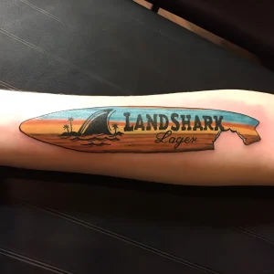 Татуировка в виде доски для серфа с акулой и надписью 'Landshark Lager'