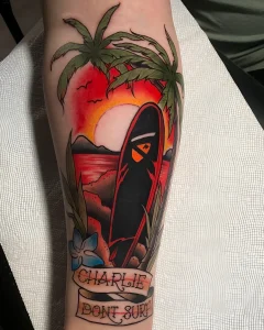 Татуировка доска для серфа среди пальм на фоне заката, с надписью 'CHARLIE DONT SURF'