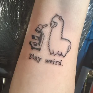 Татуировка микроскопа и милой альпаки с надписью 'Stay weird.' на предплечье.