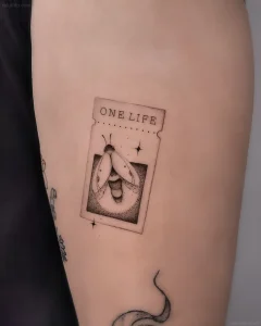 Татуировка в виде светлячка на билете с надписью "ONE LIFE", выполненная в стиле точечной графики