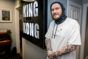 Калум Томпсон, тату-мастер из King Kong Tattoo Company, стоит в студии рядом с логотипом "KING KONG".