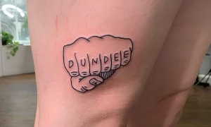 Татуировка в виде сжатого кулака с надписью DUNDEE на пальцах, выполненная в стиле традиционной татуировки.