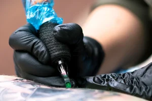 Крупный план руки мастера в черной перчатке, держащей тату-машинку, наносящей рисунок на кожу
