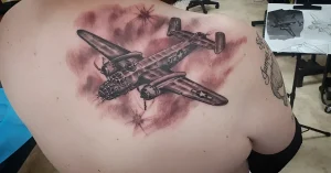 Реалистичное тату бомбардировщика B-25 Митчелл на плече, выполненное в монохромной гамме с красными акцентами, изображающее самолет в облаках и на фоне взрывов.