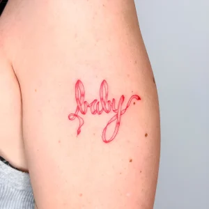 Красная акварельная татуировка слова "baby" на плече, имитирующая ленту