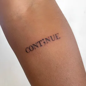 Крупный план татуировки с надписью "CONT;NUE" на предплечье, выполненной черными чернилами в минималистичном стиле.