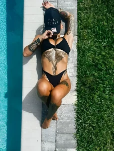 Женщина в купальнике и кепке с надписью "Fuel Girls" лежит у бассейна, демонстрируя татуировки на груди, животе, руках и ногах.