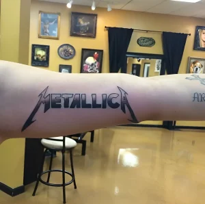 Крупный план татуировки логотипа группы Metallica на руке, выполненной в черном цвете с объемным эффектом.