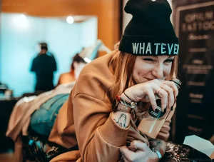 Девушка в черной шапке с надписью "WHATEVER" пьет напиток из бутылки, на ее руке видна татуировка, в фоновом режиме идет процесс татуировки.
