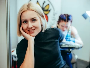 Улыбающаяся женщина с светлыми волосами в черном водолазке, рука подперта подбородок. На заднем плане размытый силуэт тату-мастера, работающего над клиентом.