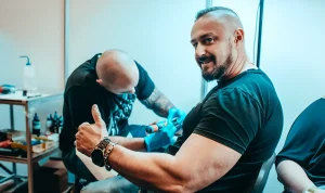 Два тату-мастера работают на Wrocław Tattoo Show 2026, один из них позирует с поднятым вверх большим пальцем.