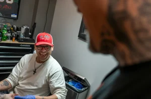 Мужчина в красной кепке с надписью "NO JOKE Tattoo Studio" смеется, пока другой мужчина делает ему татуировку.