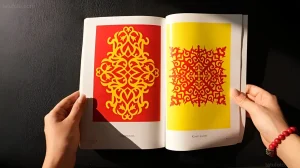 Две руки держат открытую книгу с яркими страницами, на которых изображены казахские орнаменты в стиле трафаретной печати.