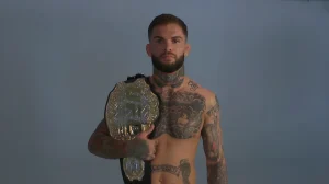 Боец UFC Коди Гарбрандт стоит, держа в руках чемпионский пояс, с видимыми татуировками на груди, шее и руках.