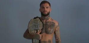 Боец UFC Коди Гарбрандт стоит, держа чемпионский пояс UFC на плече, демонстрируя многочисленные татуировки на груди, шее и руках.