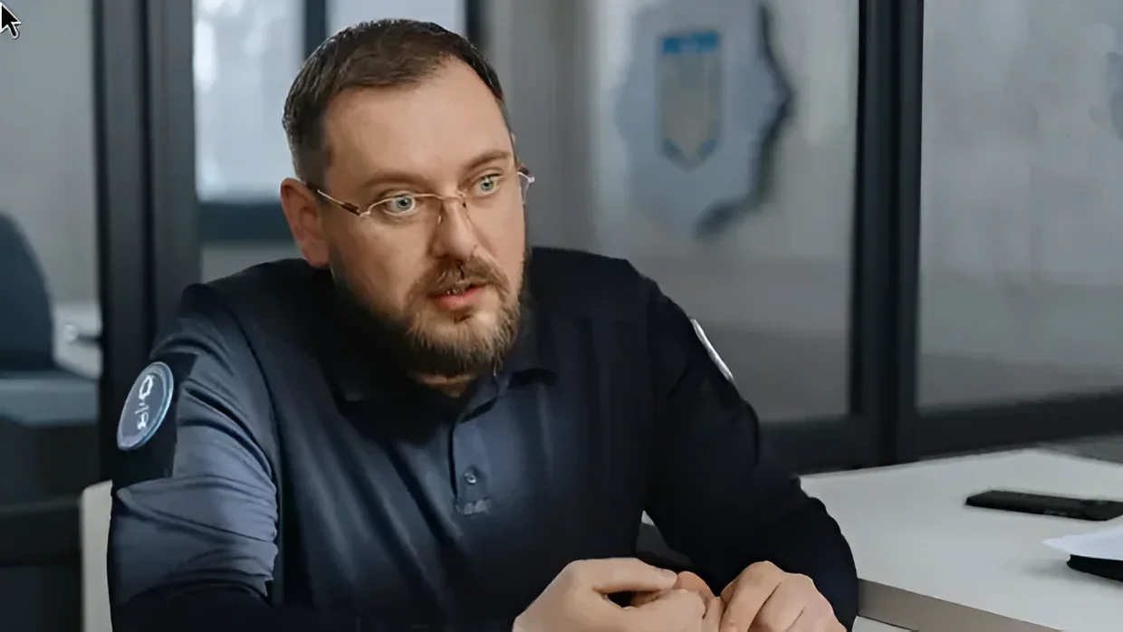 Мужчина в очках и темно-синей рубашке с нашивками сидит за столом, разговаривая.