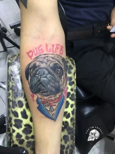 Реалистичная татуировка мопса в костюме с надписью "PUG LIFE" на предплечье