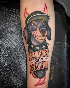 Татуировка таксы в зеленой кепке, облизывающей нос, с надписью "All cats are beautiful" и красными рожками.