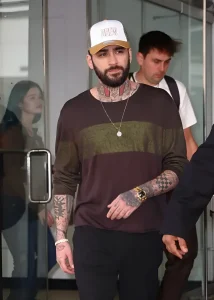 Зейн Малик в бейсболке с надписью "DREW" и в свободной кофте с горизонтальными полосами, демонстрирующий свои многочисленные татуировки на шее и руках.
